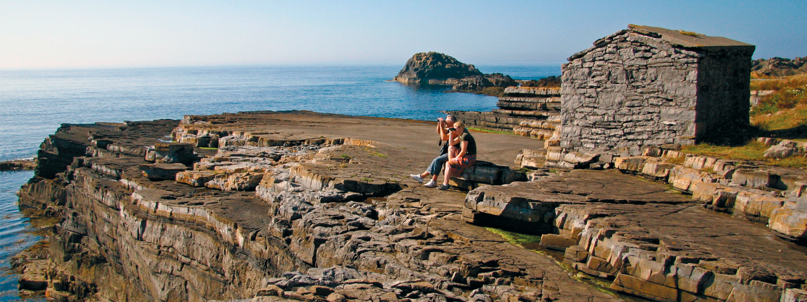 A couple sat on some rocks at Scarlett with binoculars looking through binoculars at nearby wildlife on the Isle of Man
